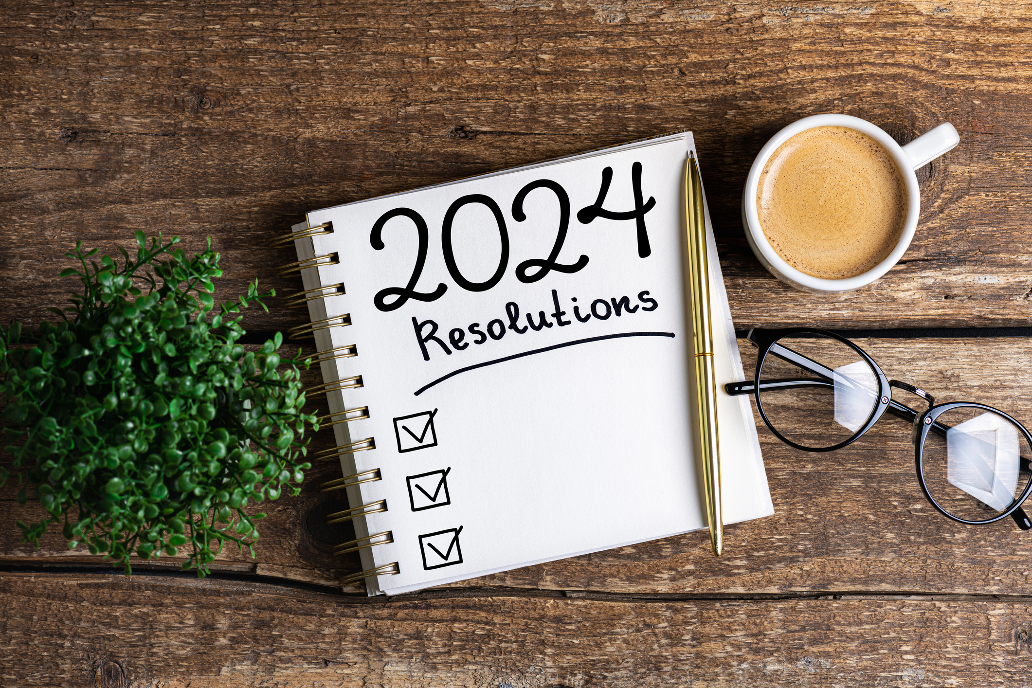 A notepad with the words “2024 Resolutions” set on a dark wood table with a coffee, pen, glasses, and plant. 